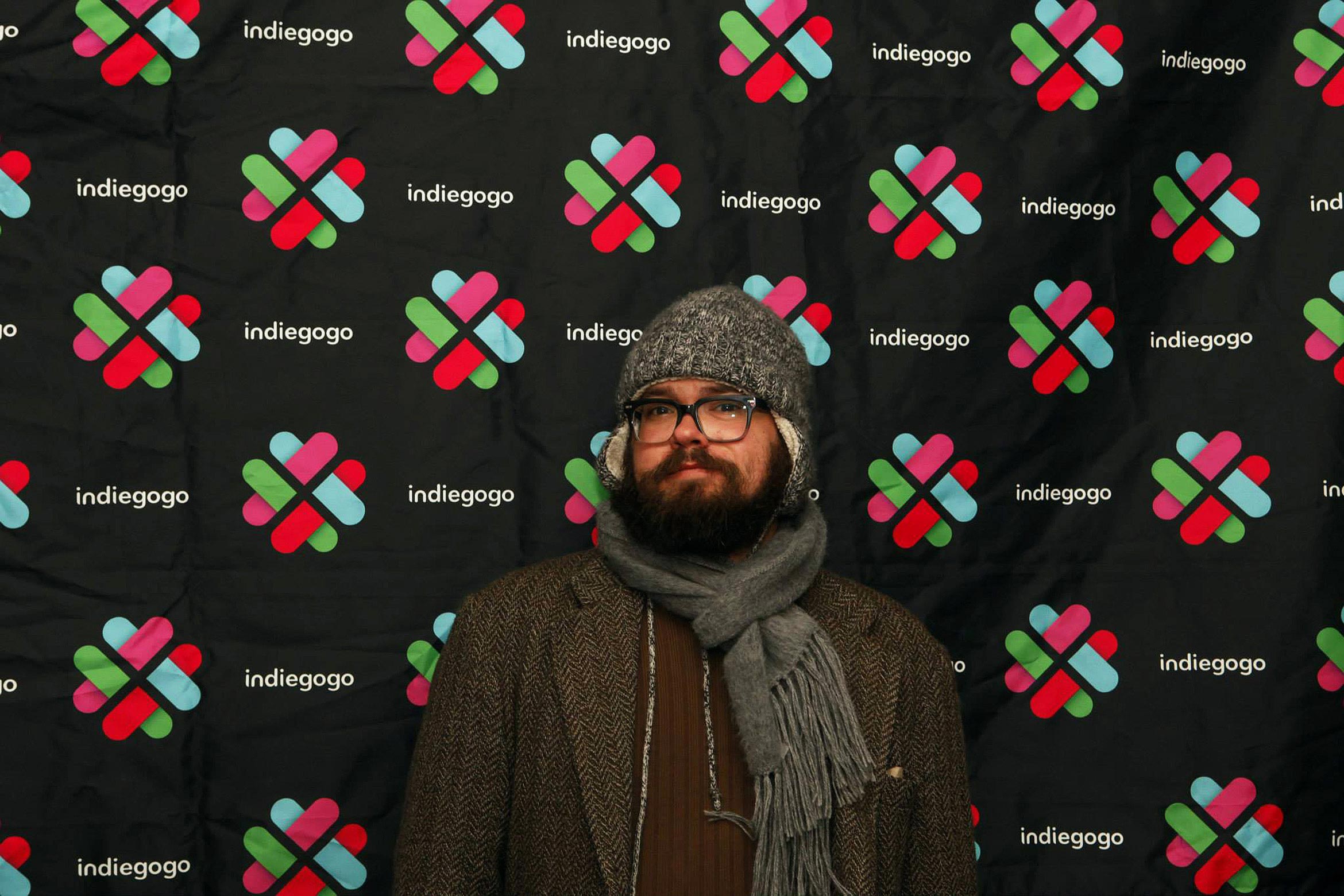 step and repeat of the indiegogo logo at the sundance music festival with bearded guy in hat, scarf and coat in front of it