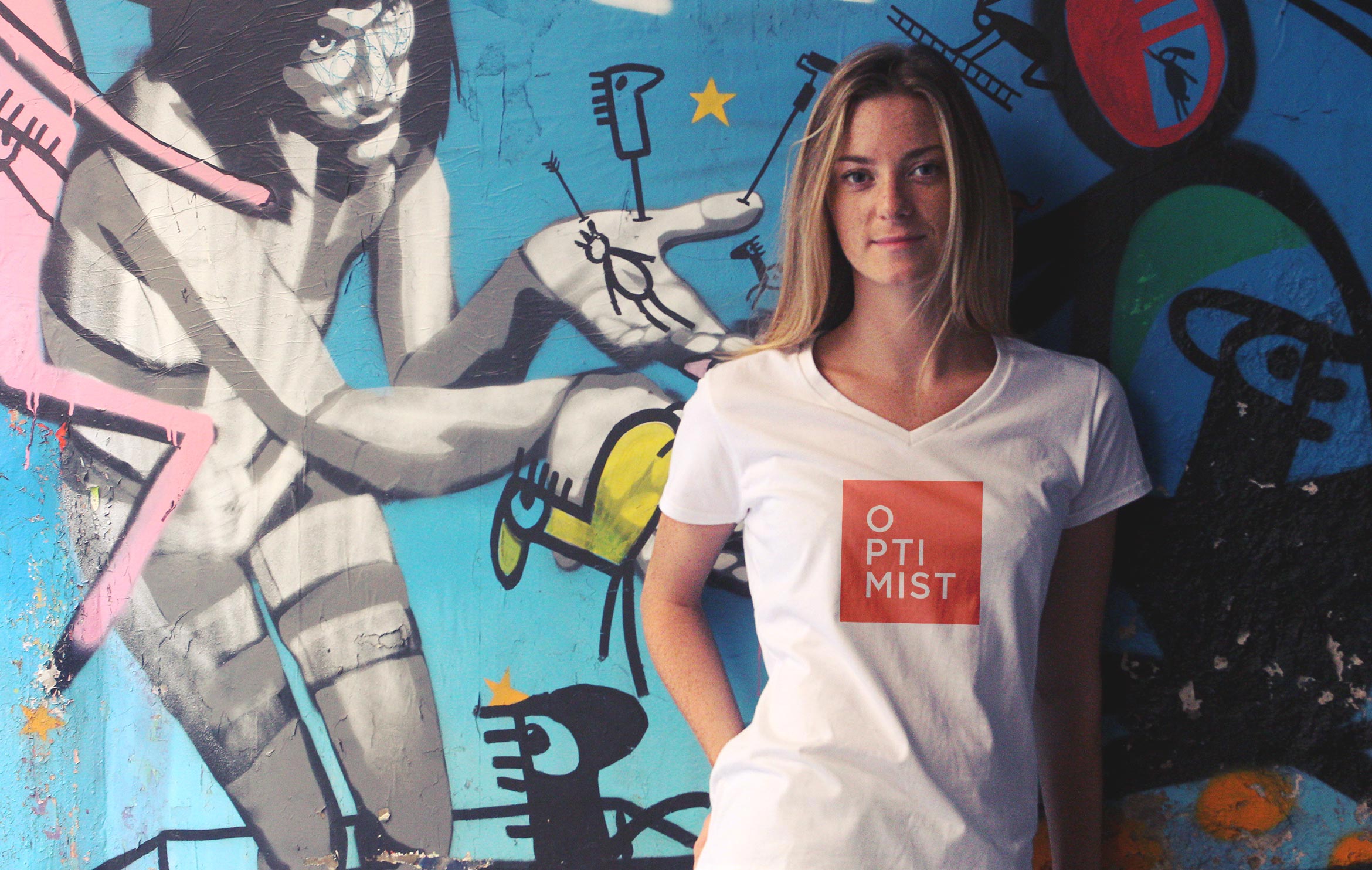 young blond woman wearing a white tee shirt with orange optimist logo in front of a graffiti wall
