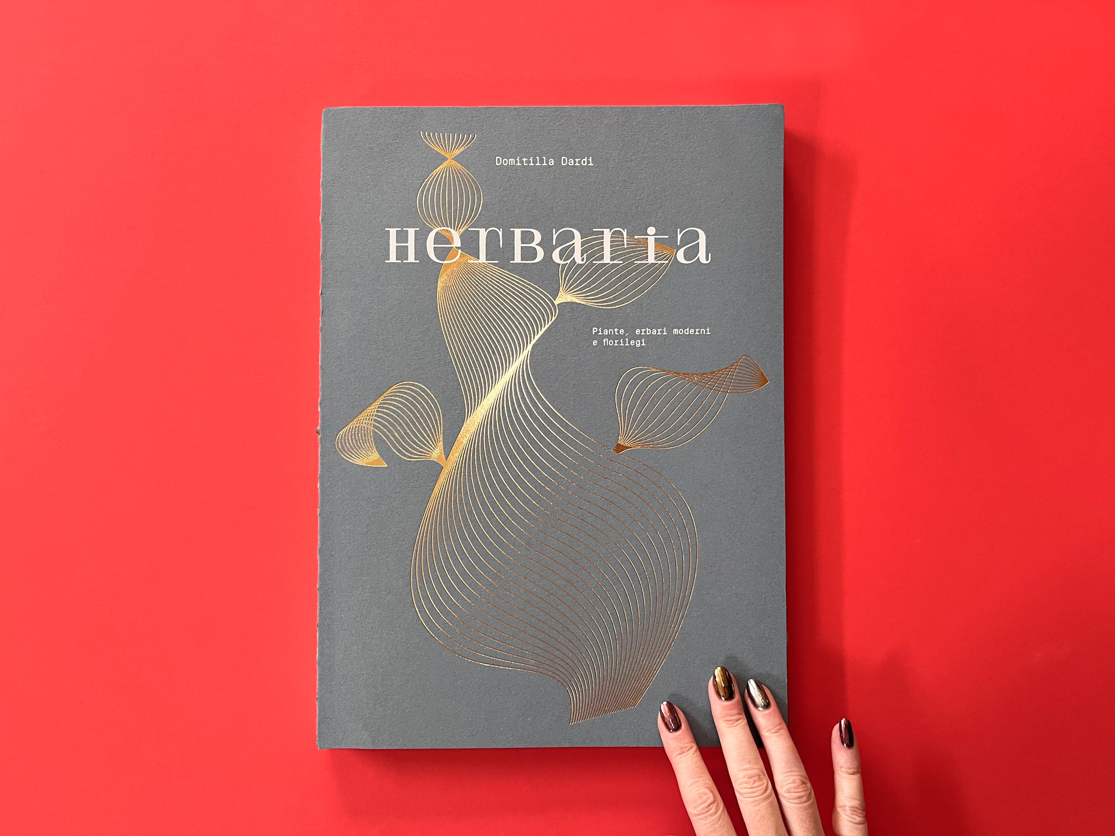 hand with metallic nail polish holding gold foil embossed herbaria magazine on red backdrop