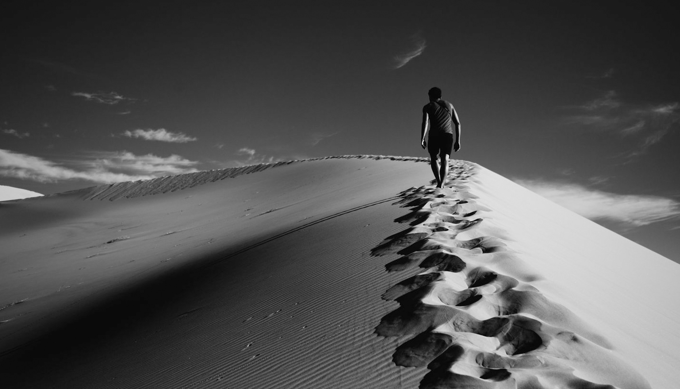 black and white image in desert with man walking at the peak of a sand hill leaving footprints