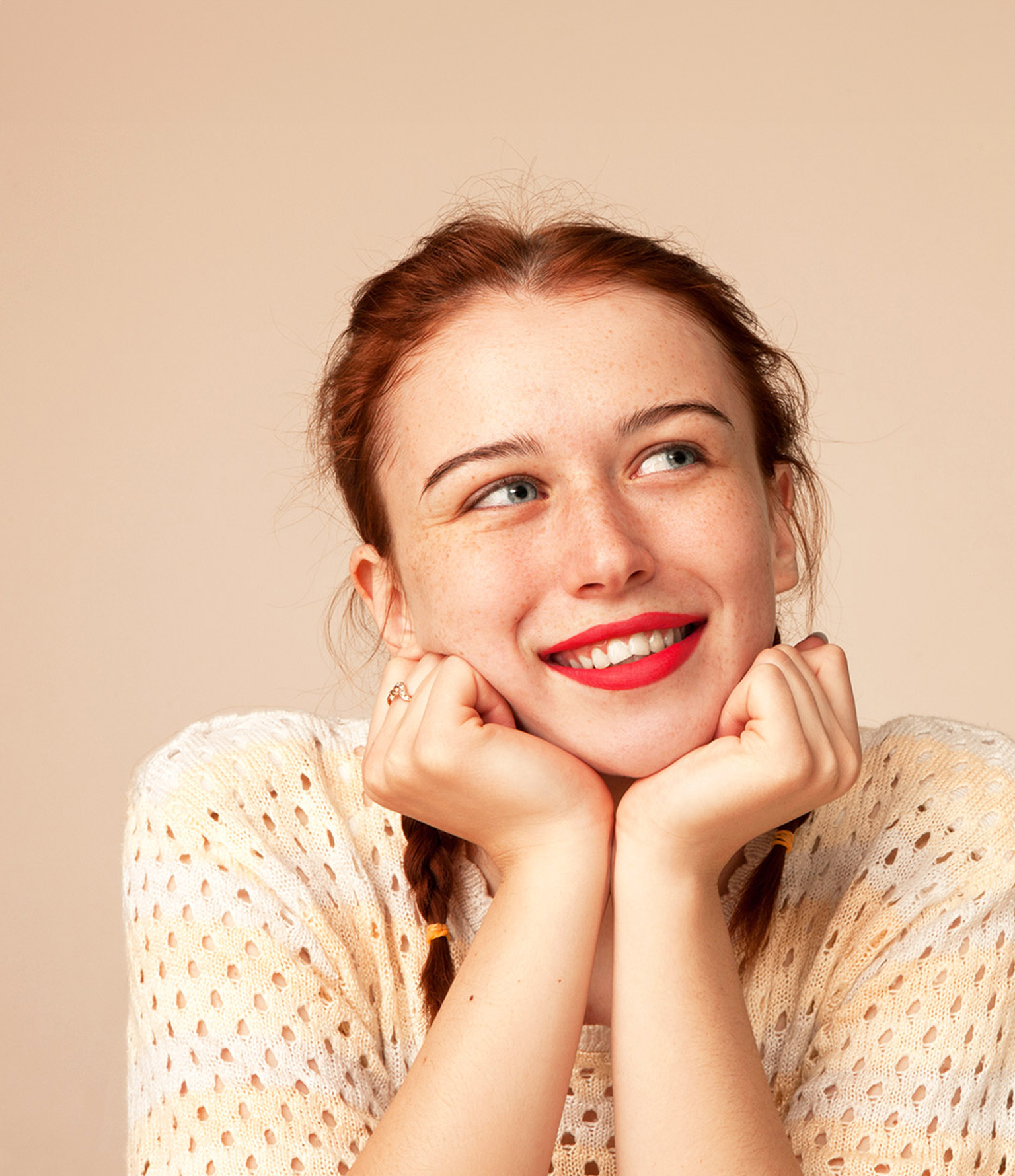 teenage girl with red lipstick and red hair smiling with head in hands on peach backdrop